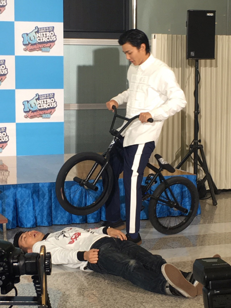 仰向けになった綾部の直前で自転車を止めておどける野村